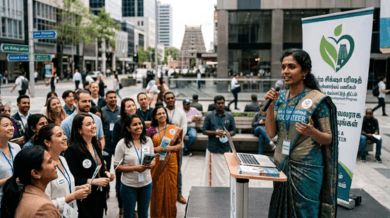A volunteer speaking to a gathered crowd at a public awareness program beside an organization banner